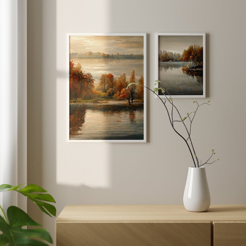 May include: Two framed landscape paintings of a lake with trees in autumn colors. The larger painting is on the left and shows a wider view of the lake. The smaller painting is on the right and shows a closer view of the trees. Both paintings have white frames.