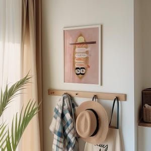 May include: A framed illustration of sushi on a pink background. The illustration features a variety of sushi rolls, including shrimp and cucumber, and a pair of chopsticks. The frame is white and has a simple design.