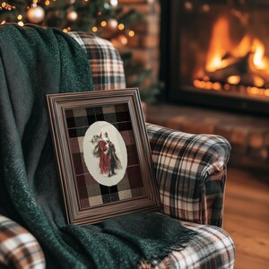 May include: A framed vintage print featuring a couple in a red and brown coat, set against a plaid background. The frame is brown, and the print is resting on a plaid armchair with a green blanket. A Christmas tree and fireplace are in the background.