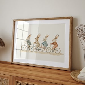 Op de afbeelding: Een ingelijste aquarelprint met vier konijnen op fietsen. Elk konijn draagt een jas in een andere kleur: grijs, beige, blauw en oranje. Het kunstwerk is geplaatst tegen een neutrale achtergrond en ingelijst in een lichtbruine houten lijst.