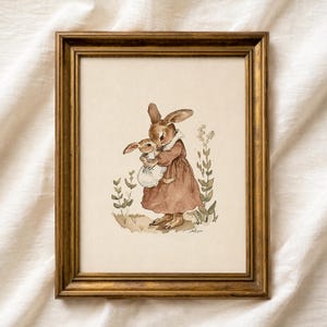 Puede incluir: Una impresión en acuarela enmarcada con dos conejos. El conejo más grande, con un vestido marrón, sostiene a un conejo más pequeño con un vestido blanco. La obra de arte tiene una estética vintage, con colores suaves y detalles delicados. El marco es dorado.