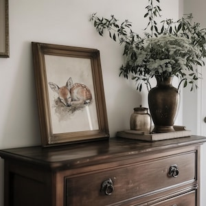 May include: A framed watercolour painting of a sleeping fawn with brown fur and white spots. The painting is in a gold frame and is hanging on a wall above a wooden dresser.