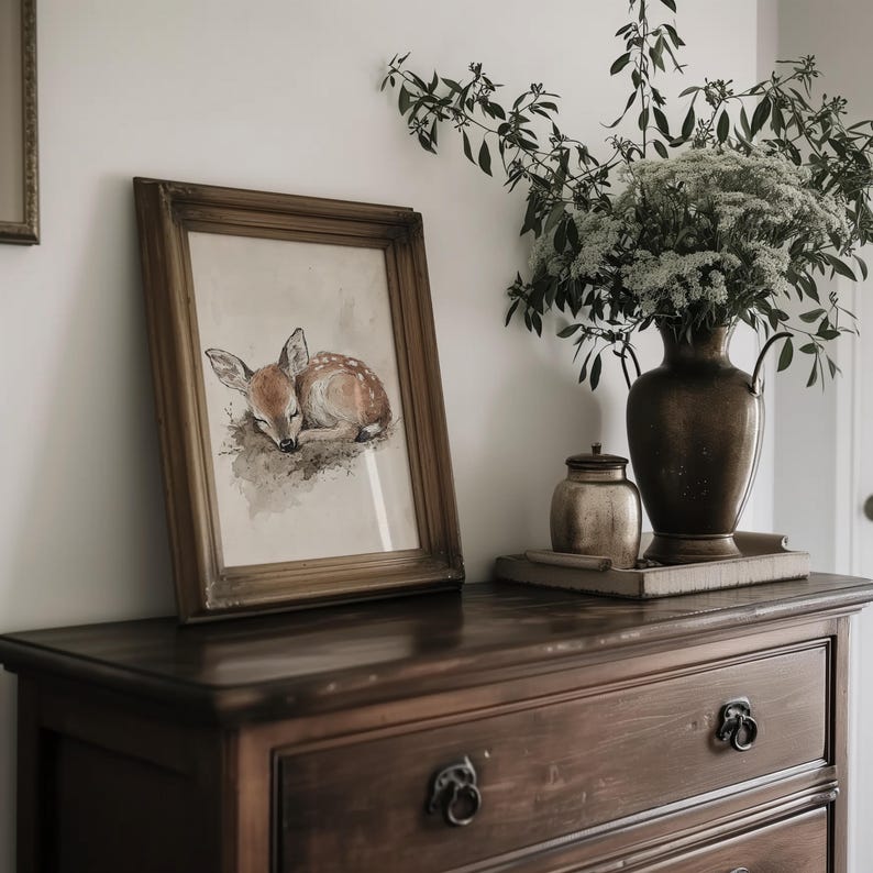 May include: A framed watercolour painting of a sleeping fawn with brown fur and white spots. The painting is in a gold frame and is hanging on a wall above a wooden dresser.