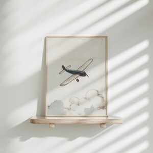 May include: A framed print featuring a vintage airplane illustration. The artwork depicts a biplane in flight, rendered in muted blue and beige tones against a cloudy background. The print is displayed on a wooden shelf.