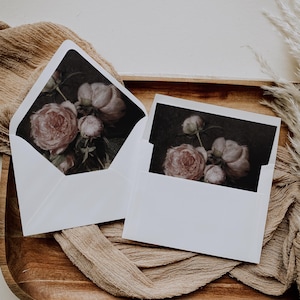 May include: Two white envelopes with floral designs. The envelopes are on a brown surface with a beige fabric underneath. The floral designs are of pink peonies.