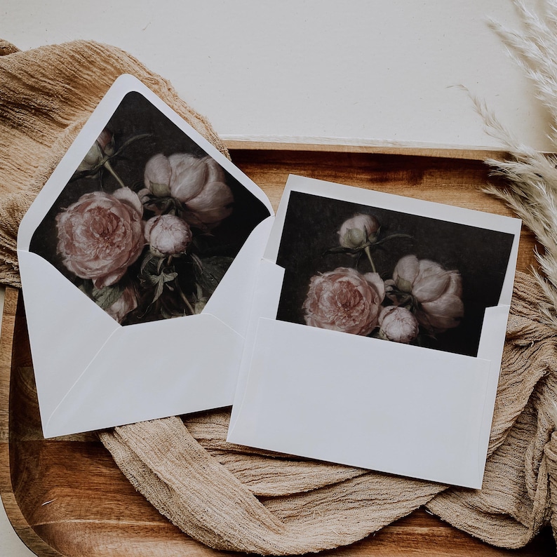 May include: Two white envelopes with floral designs. The envelopes are on a brown surface with a beige fabric underneath. The floral designs are of pink peonies.