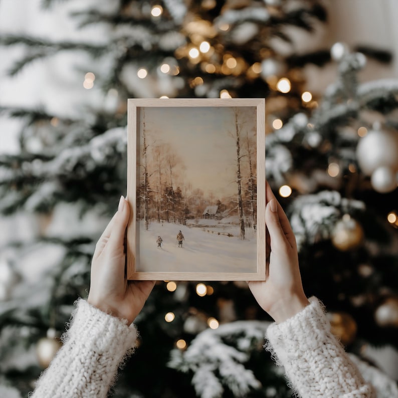 Puede incluir: Una pintura enmarcada de un paisaje invernal nevado con figuras caminando hacia una casa distante. La obra de arte est&aacute; en un marco de madera clara, sobre un fondo de un &aacute;rbol de Navidad decorado con luces suaves y borrosas.