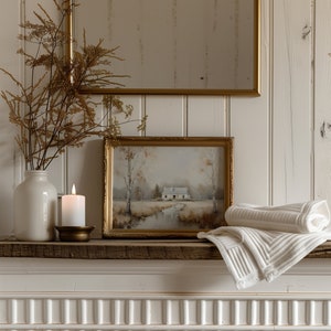 May include: A white ceramic vase with dried flowers sits on a wooden mantel with a gold candle holder and a lit candle. A framed watercolor painting of a house in a forest hangs above the vase. A white towel is draped over the mantel.
