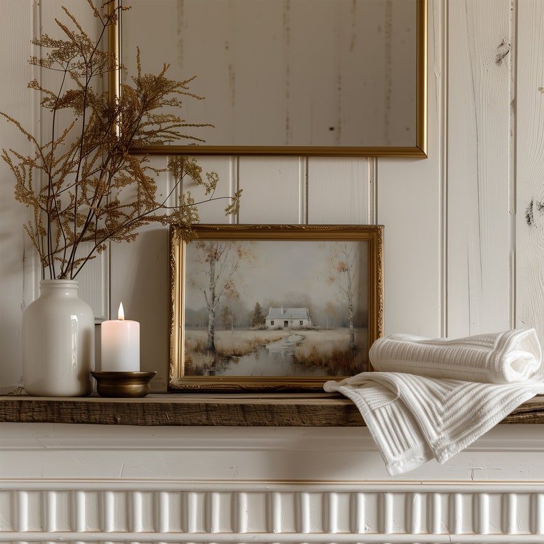 May include: A white ceramic vase with dried flowers sits on a wooden mantel with a gold candle holder and a lit candle. A framed watercolor painting of a house in a forest hangs above the vase. A white towel is draped over the mantel.