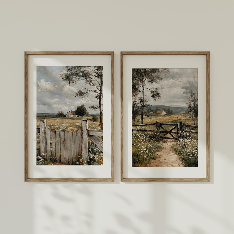 May include: Two framed landscape prints. Each print depicts a pastoral scene with a wooden fence, a path, and a house in the distance. The artwork features a field of wildflowers under a cloudy sky. The frames are a light brown color.