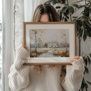 May include: A woman holds a framed painting of a cottage in a rural landscape. The painting is done in muted tones of brown, green, and blue, with a hint of white for the cottage. The frame is a simple, gold-toned wood.