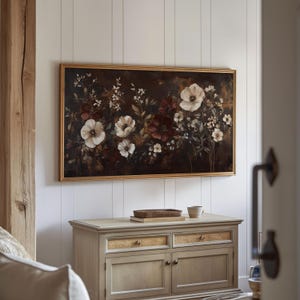 May include: A framed painting of a floral arrangement with white and red flowers on a dark background. The painting is hanging on a white wall above a light brown wooden cabinet with two drawers and two doors.