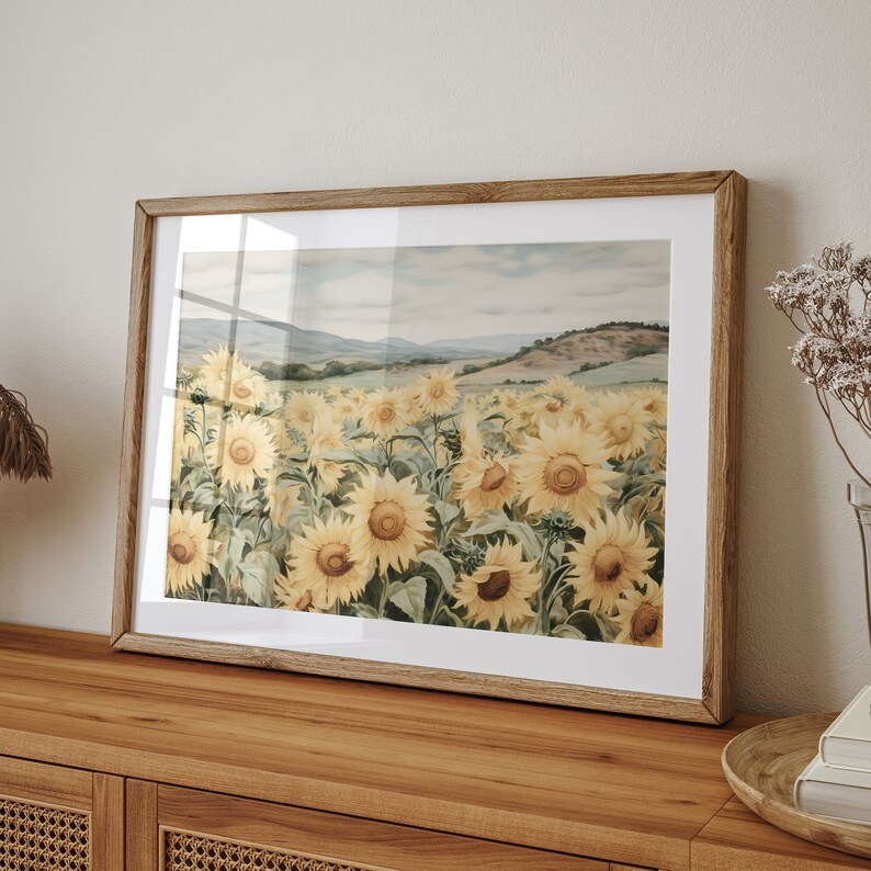 May include: A framed print of a watercolour painting depicting a field of sunflowers in bloom. The sunflowers are yellow with brown centres and green leaves. The background is a soft blue sky with a distant landscape.