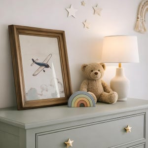 May include: A framed print of a blue airplane flying above clouds, a teddy bear, a rainbow-shaped wooden toy, and a white lamp with a beige lampshade sit on a sage green dresser. Star-shaped drawer pulls and wall decorations complete the nursery decor.