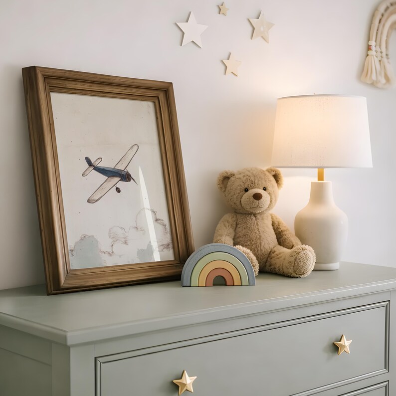 May include: A framed print of a blue airplane flying above clouds, a teddy bear, a rainbow-shaped wooden toy, and a white lamp with a beige lampshade sit on a sage green dresser. Star-shaped drawer pulls and wall decorations complete the nursery decor.