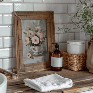May include: A framed painting of a bouquet of pink roses in a vintage teacup, with a white towel, a brown glass bottle with a pump dispenser, and a white ceramic jar on a rustic wooden surface.