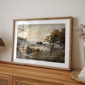 May include: A framed watercolor painting depicting a landscape scene with a lake, trees, and hills in the distance. The painting is done in shades of brown and gray, with a soft, muted color palette.