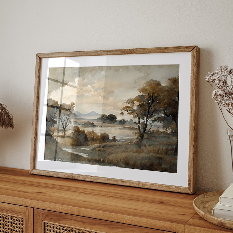 May include: A framed watercolor painting depicting a landscape scene with a lake, trees, and hills in the distance. The painting is done in shades of brown and gray, with a soft, muted color palette.