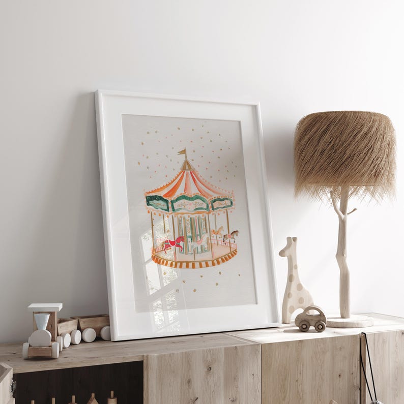 May include: A white framed print of a watercolour painting of a carousel with a red, orange and green striped canopy. The carousel is surrounded by gold stars on a light grey background.