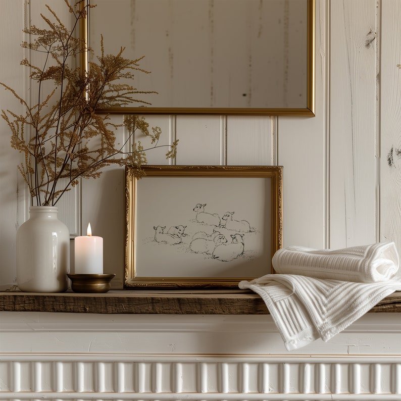 May include: A white ceramic vase with dried flowers sits on a wooden shelf with a gold candle holder and a framed black and white drawing of a group of lambs. Two white towels are folded on the shelf.