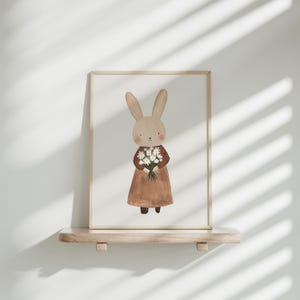 May include: A framed illustration of a brown rabbit wearing a brown dress and holding a bouquet of white flowers.