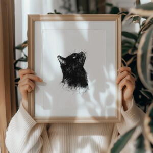 May include: A black and white line drawing of a cat looking up, framed in a light brown wood frame.