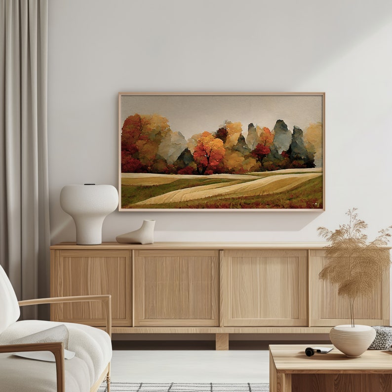 May include: A framed landscape painting depicting a field with trees in the background. The trees are a mixture of green, brown, and orange. The field is a light brown colour. The painting is in a wooden frame.