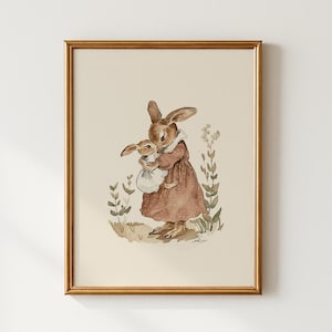 May include: A framed watercolour print depicting two brown rabbits. One larger rabbit in a brown dress holds a smaller rabbit in a white dress. The artwork has a vintage aesthetic with soft colours and botanical accents.