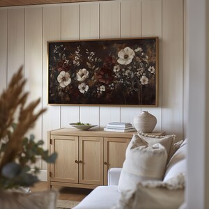 May include: A framed floral painting with a dark background and white and burgundy flowers. The painting is hanging on a light wood wall above a light wood cabinet with three doors. A white couch with a white throw pillow is in the foreground.