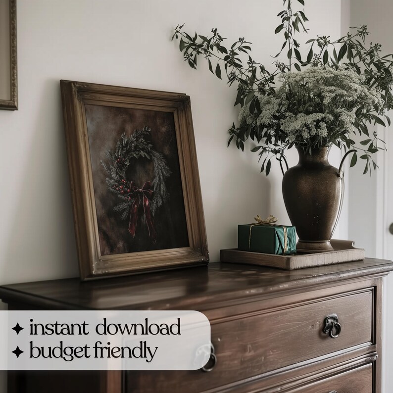 Op de afbeelding: Een ingelijste print van een donkergroene krans met rode bessen en een rood lint op een bruine achtergrond. De print hangt aan een donkerhouten dressoir met een groene cadeaudoos erop.