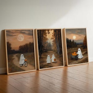 May include: Three framed art prints depicting ghosts in an autumnal setting. One rides a bicycle with a pumpkin, others walk with jack-o'-lanterns or push a cart of pumpkins. The artwork uses warm colours, creating a cosy atmosphere, perfect for Halloween.