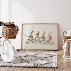 Op de afbeelding: Ingelijste aquarelprint van vier konijnen op fietsen, gekleed in verschillende outfits. Een witte driewieler met een rieten mand staat op de voorgrond. Een grijs tapijt en een kleine houten tafel zijn ook zichtbaar, in een kinderkamer.