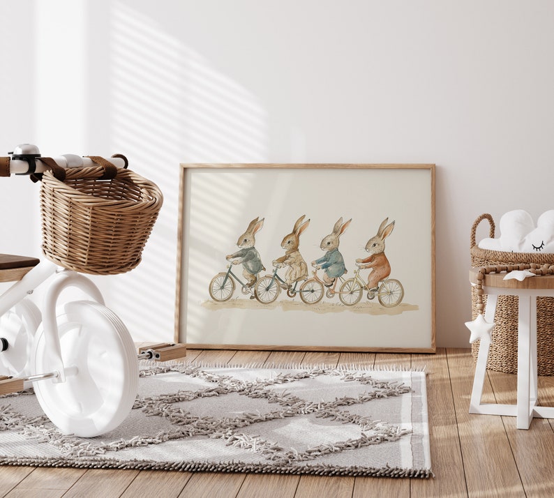 Op de afbeelding: Ingelijste aquarelprint van vier konijnen op fietsen, gekleed in verschillende outfits. Een witte driewieler met een rieten mand staat op de voorgrond. Een grijs tapijt en een kleine houten tafel zijn ook zichtbaar, in een kinderkamer.