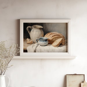 Peut inclure: Une peinture encadr&eacute;e repr&eacute;sente une nature morte avec un pain, une tranche et un petit pichet bleu et blanc. Un grand pichet blanc &agrave; rayures bleues et un tissu blanc compl&egrave;tent la composition. L'&oelig;uvre a une esth&eacute;tique vintage et rustique.