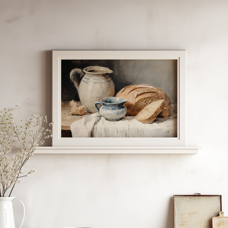 Peut inclure: Une peinture encadr&eacute;e repr&eacute;sente une nature morte avec un pain, une tranche et un petit pichet bleu et blanc. Un grand pichet blanc &agrave; rayures bleues et un tissu blanc compl&egrave;tent la composition. L'&oelig;uvre a une esth&eacute;tique vintage et rustique.