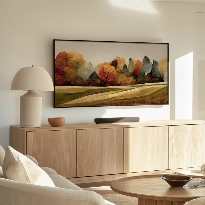 May include: A living room interior with a large framed painting of a forest scene with autumn foliage hanging on a wall above a wooden cabinet. A lamp sits on the cabinet next to a small bowl. A coffee table with a bowl and a magazine sits in front of a white sofa.