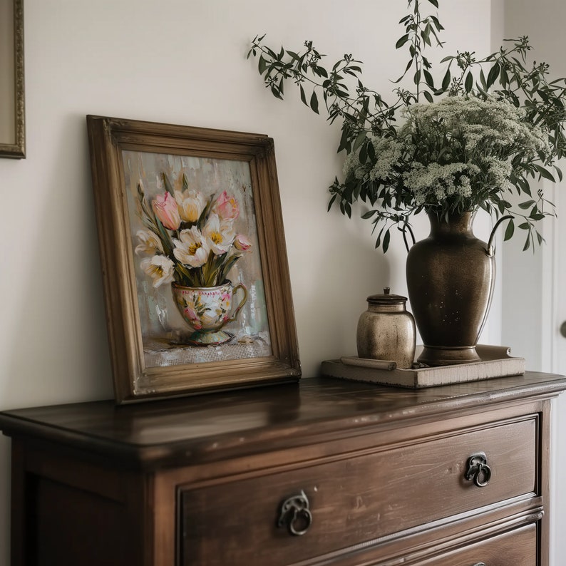 May include: A framed painting of a bouquet of pink and white tulips in a vintage teacup, set on a wooden dresser with a vase of white flowers and a small decorative jar.