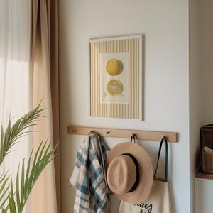 May include: A white framed print with a yellow and white striped border. The print features a drawing of a whole and a sliced citrus fruit. The print is hanging on a wall above a wooden coat rack with a plaid scarf, a straw hat, and a tote bag hanging on it.