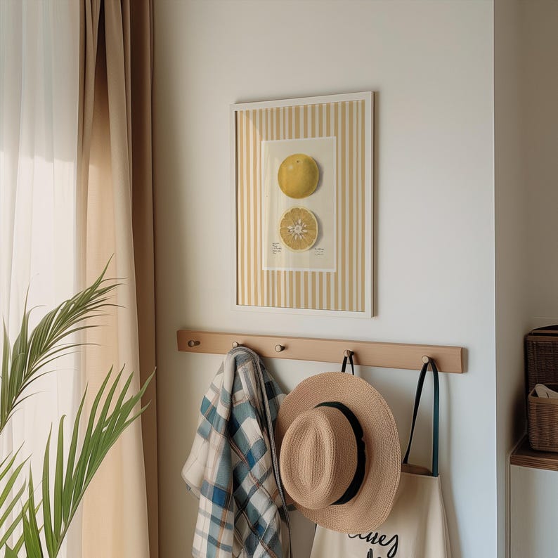 May include: A white framed print with a yellow and white striped border. The print features a drawing of a whole and a sliced citrus fruit. The print is hanging on a wall above a wooden coat rack with a plaid scarf, a straw hat, and a tote bag hanging on it.