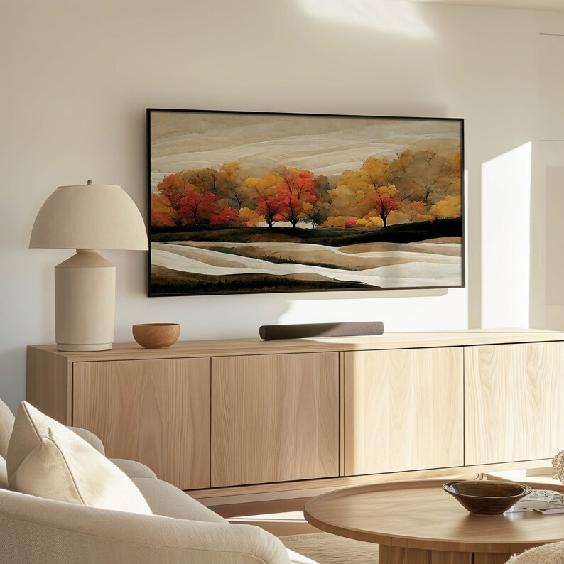 Puede incluir: Un armario de madera clara con tres puertas y una barra de sonido debajo. Una l&aacute;mpara con una pantalla beige se encuentra a la izquierda del armario. Una pintura enmarcada de un paisaje con &aacute;rboles en colores oto&ntilde;ales cuelga sobre el armario.