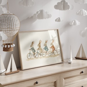 Op de afbeelding: Een ingelijste aquarelprint met vier konijnen op fietsen. De konijnen dragen jassen in grijs, beige, blauw en oranje. De print staat op een lichte houten dressoir, met decoratieve wolken en een heteluchtballon op de achtergrond.