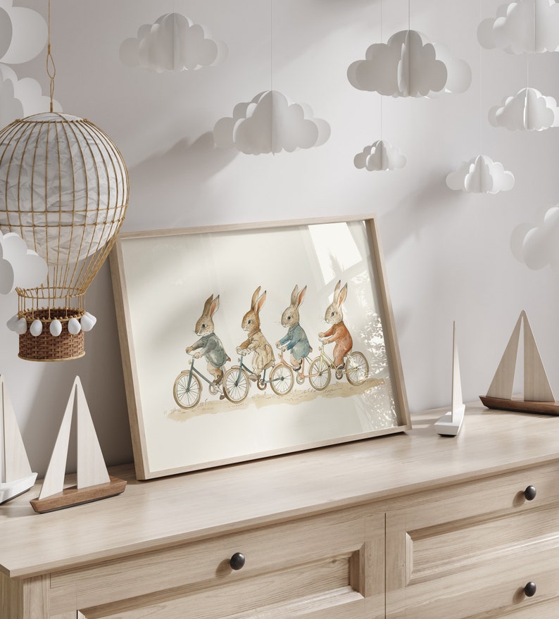 Op de afbeelding: Een ingelijste aquarelprint met vier konijnen op fietsen. De konijnen dragen jassen in grijs, beige, blauw en oranje. De print staat op een lichte houten dressoir, met decoratieve wolken en een heteluchtballon op de achtergrond.