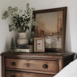 Puede incluir: Una c&oacute;moda de madera con una pintura enmarcada de un paisaje invernal y una peque&ntilde;a imagen enmarcada con un dise&ntilde;o de reno. Un jarr&oacute;n de flores, un peque&ntilde;o &aacute;rbol de Navidad y libros tambi&eacute;n est&aacute;n en la c&oacute;moda.