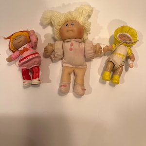 Puede incluir: Tres muñecas Cabbage Patch Kids, una con cabello rubio y un vestido rosa, otra con cabello naranja y un vestido rosa, y la última con cabello amarillo y un vestido amarillo.