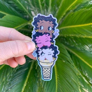 May include: A sticker of three cartoon faces stacked on top of each other in an ice cream cone. The top face is brown with black hair and sprinkles, the middle face is pink with black hair, and the bottom face is white with black hair and a wink. The cone is yellow and white with the text "SLIM B DESIGNS" on the side.