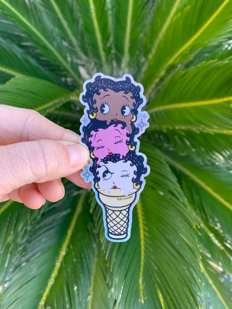May include: A sticker of three cartoon faces stacked on top of each other in an ice cream cone. The top face is brown with black hair and sprinkles, the middle face is pink with black hair, and the bottom face is white with black hair and a wink. The cone is yellow and white with the text "SLIM B DESIGNS" on the side.
