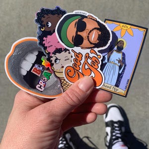 May include: A collection of colorful stickers featuring a cartoon ice cream cone, a mouth with teeth, a person with sunglasses, and a religious figure. The stickers are held in a hand wearing black and white sneakers.
