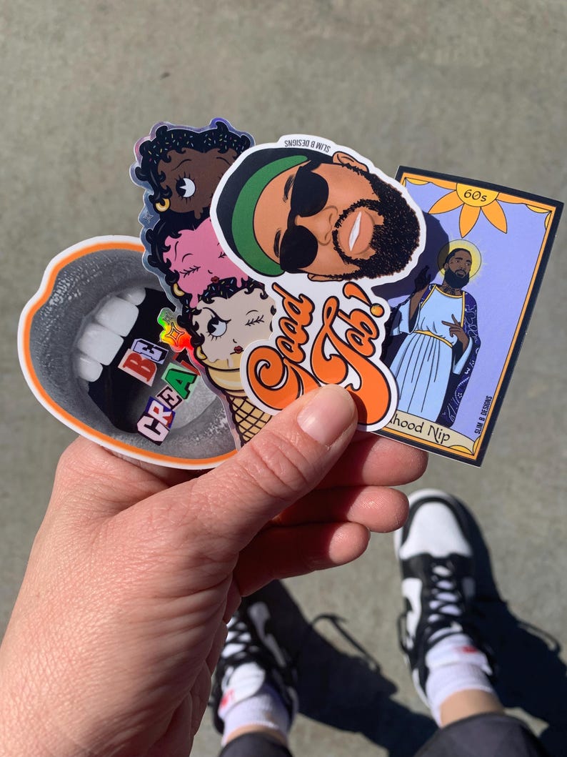 May include: A collection of colorful stickers featuring a cartoon ice cream cone, a mouth with teeth, a person with sunglasses, and a religious figure. The stickers are held in a hand wearing black and white sneakers.