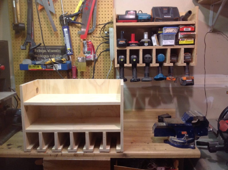 May include: A wooden storage shelf with two shelves and multiple compartments for organizing tools. The shelf is made of light-colored wood and has a simple design. It is sitting on a wooden table next to a blue metal vise.