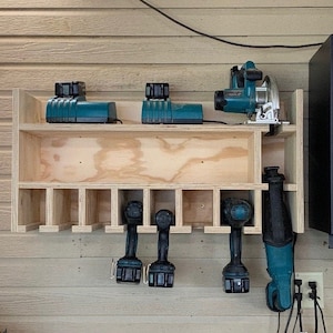 May include: A wooden wall-mounted storage rack with multiple compartments for organizing power tools. The rack features a top shelf with two teal power tool batteries and a teal circular saw.  The bottom shelf has eight compartments for storing tools, with three teal cordless drills and a teal reciprocating saw hanging from the bottom.